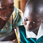 A student writes with a pencil while a teacher is nearby