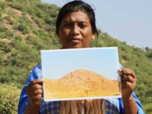 A woman standing in a lush green landscape holds a photograph of a brown landscape