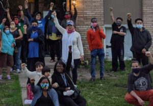 A group of people, many wearing masks, sit and stand outside and raise their fists