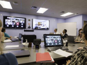A group of people in a conference room participate in a virtual meeting
