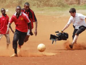 Men play football (soccer) while a man films them with a professional camera